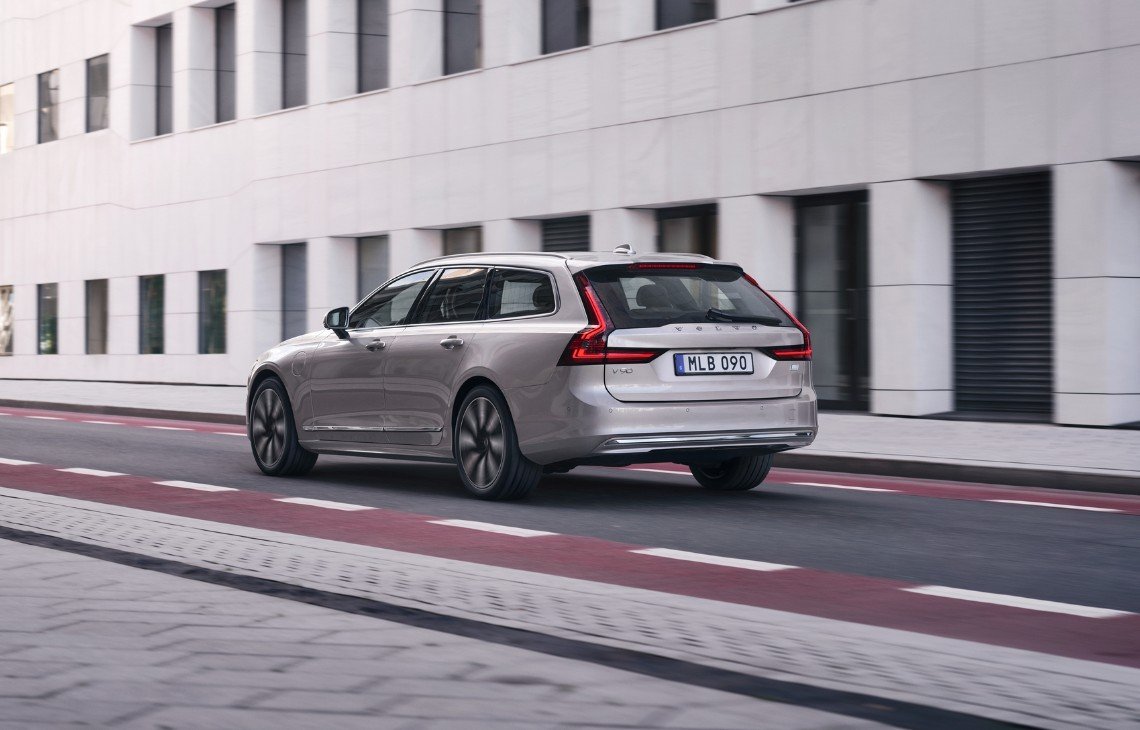 Volvo V90 INSCRIPTION in Bright Silver Metallic steht neben einem modernen, beleuchteten Haus auf einem Steinboden - Heckseitenschuss